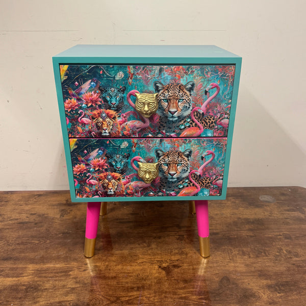 A small turquoise side table with two drawers, decorated with colorful tropical and floral artwork featuring leopard faces. The table has bright pink legs with gold metal caps, set on a wooden table against a plain wall.